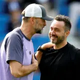Zwei Fußballtrainer, einer mit Kappe, lachen sich an. De Zerbi, der Trainer von Brighton, legt dem anderen die Hand auf die Schulter.