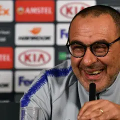 Lachender Sarri Trainer mit Brille und blauem Kragen. Hinter ihm Logos von Sponsoren.