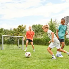 Drei Kinder üben im Garten Fußball. Ein Junge dribbelt, während ein Mädchen und ein weiterer Junge zuschauen.