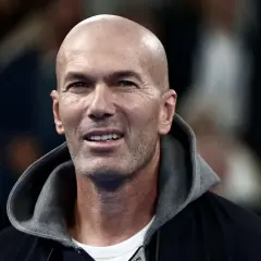 Zinedine Zidane, mit seinem charakteristischen Glatzkopf und einem Lächeln, trägt eine Kapuzenjacke. Seine Rolle als Fußballlegende ist unbestritten.