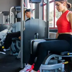 Frau trainiert ihre abduktoren muskeln an einem Gerät im Fitnessstudio. Ein Mann trainiert im Hintergrund.