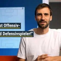 Ein Mann im weißen T-Shirt spricht über Fußballtaktiken. Er erklärt, dass jeder Spieler sowohl offensiv als auch defensiv gegen den Ball spielen muss.