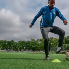 Fußballer trainiert seine motorische Schnelligkeit auf einem Kunstrasenplatz, umgeben von Bäumen und einem bewölkten Himmel.