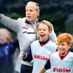 Horst Wein gibt Anweisungen, während zwei junge Fußballer mit roten Haaren und leuchtenden Augen auf dem Spielfeld jubeln.