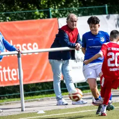Fußball-Eltern schauen ihren Kindern beim Spiel zu. Ein Junge im roten Trikot mit der Nummer 20 spielt den Ball.