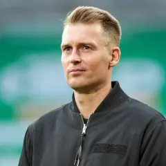 Ein Mann mit blondem Haar und blauen Augen, trägt eine schwarze Bomberjacke. Er blickt nachdenklich zur Seite.