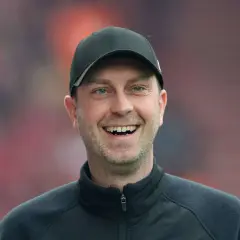 Lachender rb leipzig trainer mit schwarzer Kappe und Trainingsjacke vor unscharfem Publikum.