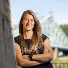 Marie-Louise Eta, lächelnd mit verschränkten Armen vor einer Brücke.