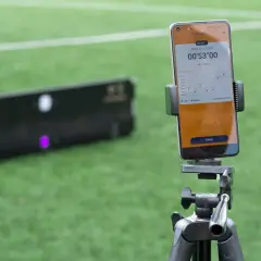 Smartphone mit Trainings-App für SAQ-Training auf einem Stativ, daneben ein Ball und ein Trainingsgerät auf einem Fußballfeld.