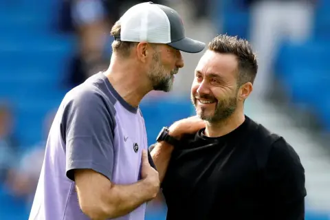 Zwei Fußballtrainer, einer mit Kappe, lachen sich an. De Zerbi, der Trainer von Brighton, legt dem anderen die Hand auf die Schulter.
