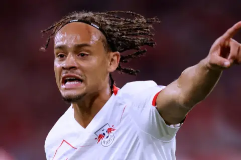 Xavi Simons, mit Dreadlocks und einem Stirnband, zeigt aufgeregt auf etwas, während er das RB Leipzig Trikot trägt.