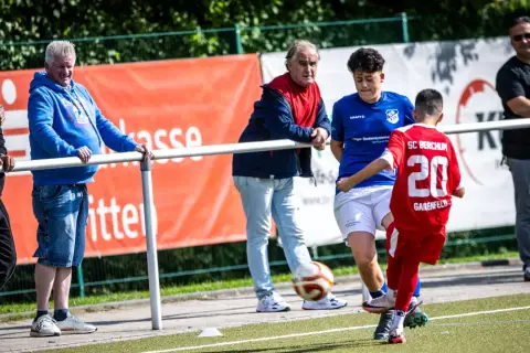 Fußball-Eltern schauen ihren Kindern beim Spiel zu. Ein Junge im roten Trikot mit der Nummer 20 spielt den Ball.