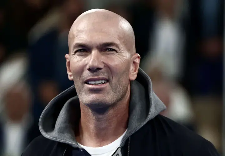 Zinedine Zidane, mit seinem charakteristischen Glatzkopf und einem Lächeln, trägt eine Kapuzenjacke. Seine Rolle als Fußballlegende ist unbestritten.