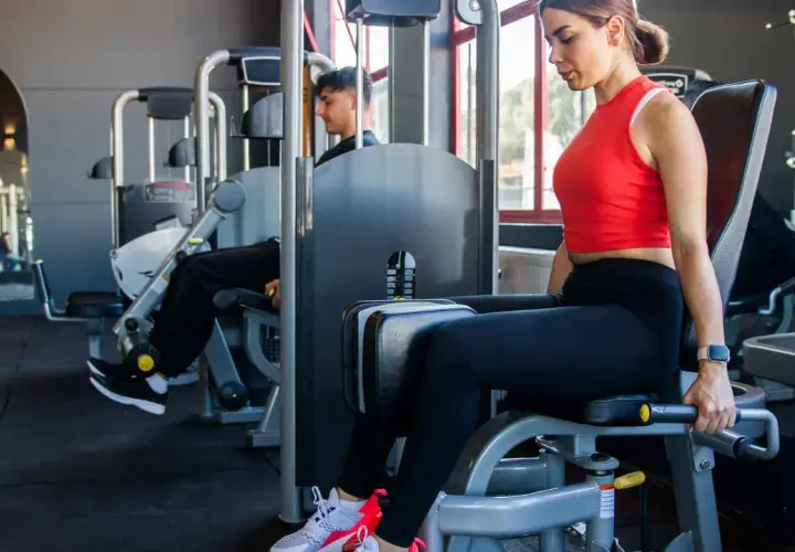 Frau trainiert ihre abduktoren muskeln an einem Gerät im Fitnessstudio. Ein Mann trainiert im Hintergrund.