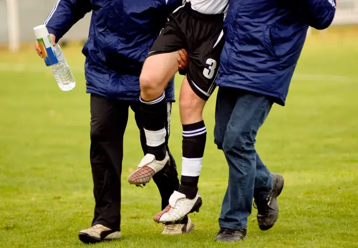 Fußballer mit verletztem Bein wird von zwei Helfern gestützt. Die Adduktoren-Gegenspieler sind hier wohl überlastet.