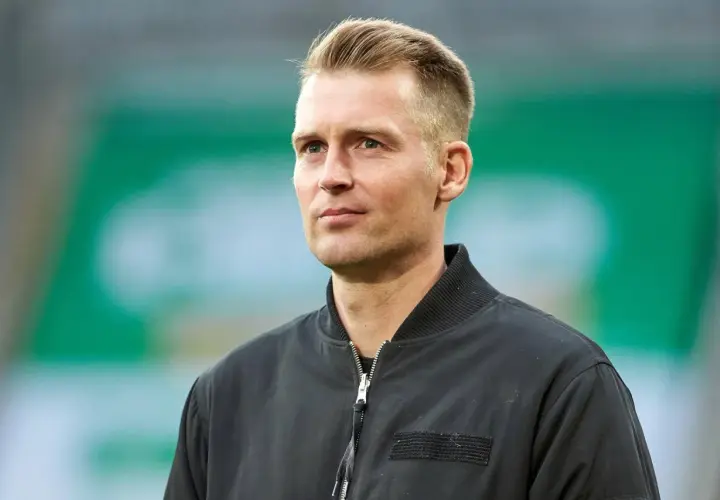 Ein Mann mit blondem Haar und blauen Augen, trägt eine schwarze Bomberjacke. Er blickt nachdenklich zur Seite.