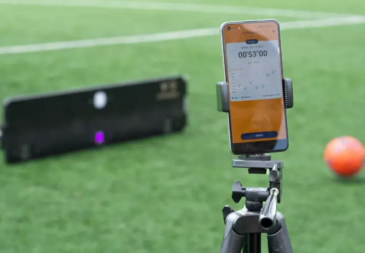 Smartphone mit Trainings-App für SAQ-Training auf einem Stativ, daneben ein Ball und ein Trainingsgerät auf einem Fußballfeld.