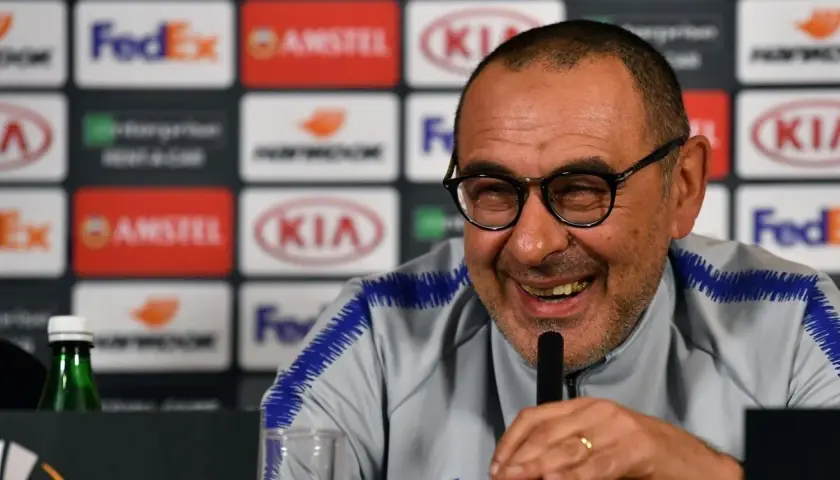 Lachender Sarri Trainer mit Brille und blauem Kragen. Hinter ihm Logos von Sponsoren.