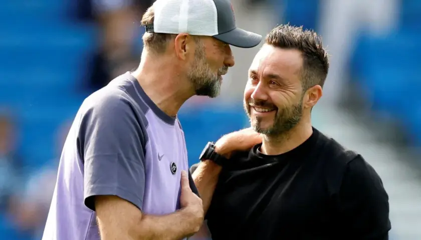 Zwei Fußballtrainer, einer mit Kappe, lachen sich an. De Zerbi, der Trainer von Brighton, legt dem anderen die Hand auf die Schulter.