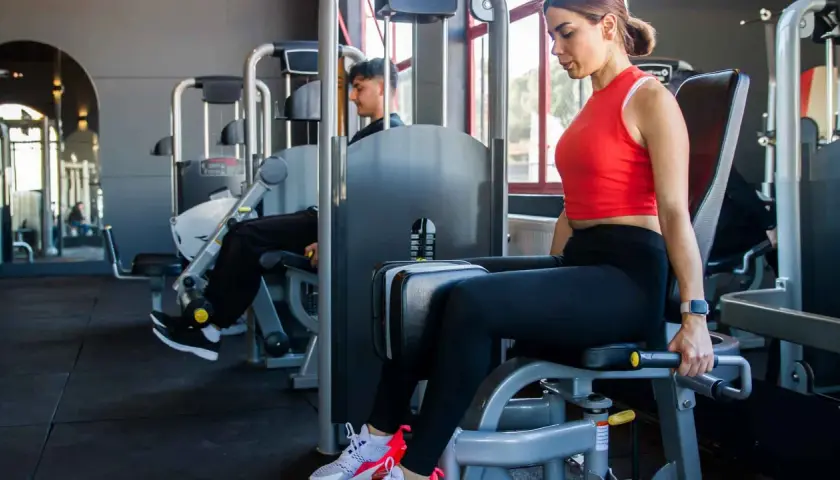 Frau trainiert ihre abduktoren muskeln an einem Gerät im Fitnessstudio. Ein Mann trainiert im Hintergrund.