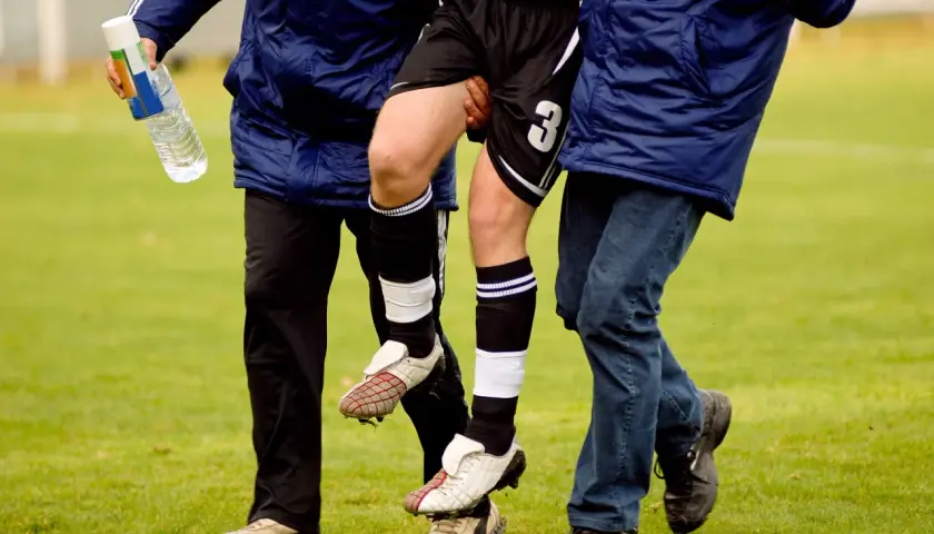 Fußballer mit verletztem Bein wird von zwei Helfern gestützt. Die Adduktoren-Gegenspieler sind hier wohl überlastet.