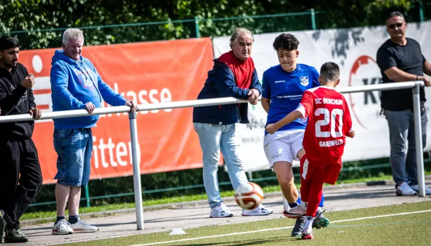 Fußball-Eltern schauen ihren Kindern beim Spiel zu. Ein Junge im roten Trikot mit der Nummer 20 spielt den Ball.