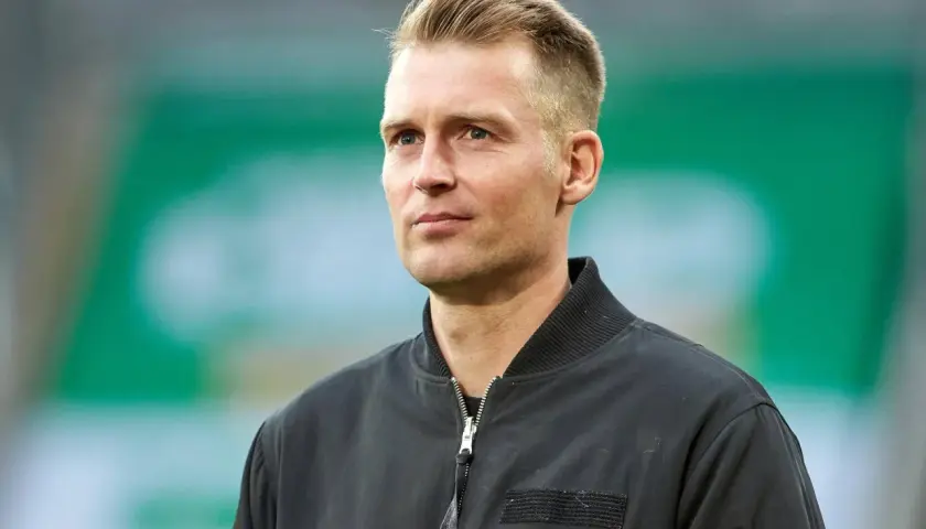 Ein Mann mit blondem Haar und blauen Augen, trägt eine schwarze Bomberjacke. Er blickt nachdenklich zur Seite.