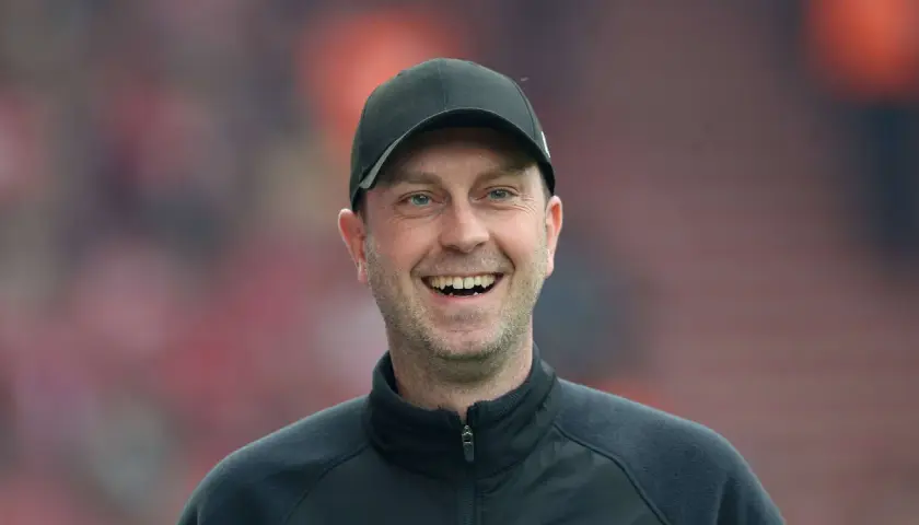 Lachender rb leipzig trainer mit schwarzer Kappe und Trainingsjacke vor unscharfem Publikum.