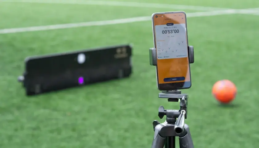 Smartphone mit Trainings-App für SAQ-Training auf einem Stativ, daneben ein Ball und ein Trainingsgerät auf einem Fußballfeld.