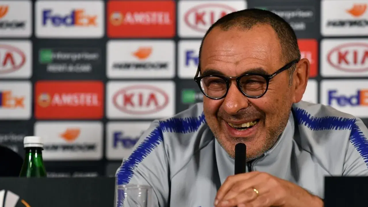 Lachender Sarri Trainer mit Brille und blauem Kragen. Hinter ihm Logos von Sponsoren.