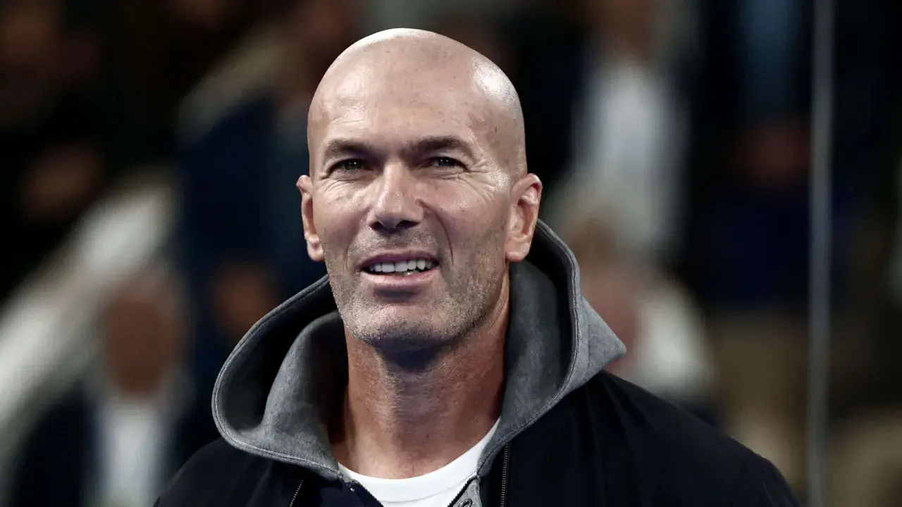 Zinedine Zidane, mit seinem charakteristischen Glatzkopf und einem Lächeln, trägt eine Kapuzenjacke. Seine Rolle als Fußballlegende ist unbestritten.