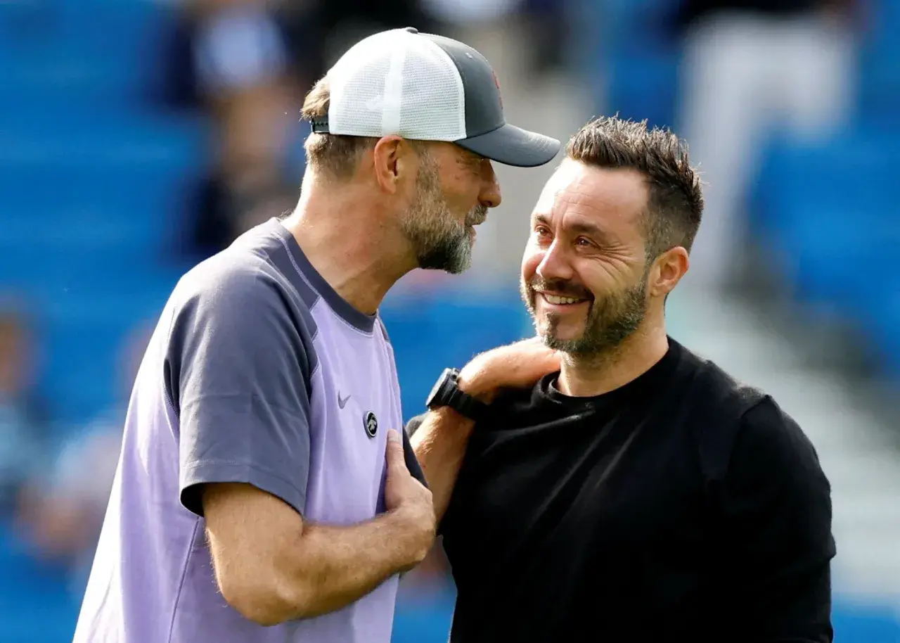 Zwei Fußballtrainer, einer mit Kappe, lachen sich an. De Zerbi, der Trainer von Brighton, legt dem anderen die Hand auf die Schulter.