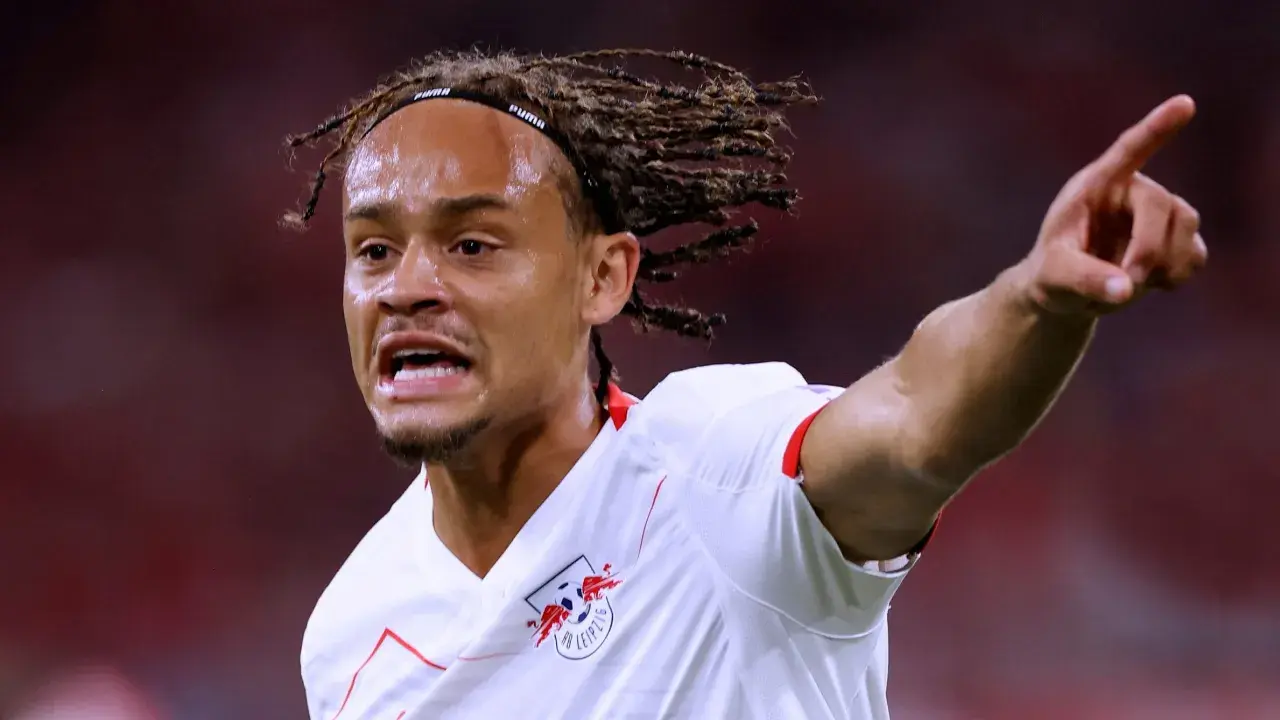 Xavi Simons, mit Dreadlocks und einem Stirnband, zeigt aufgeregt auf etwas, während er das RB Leipzig Trikot trägt.