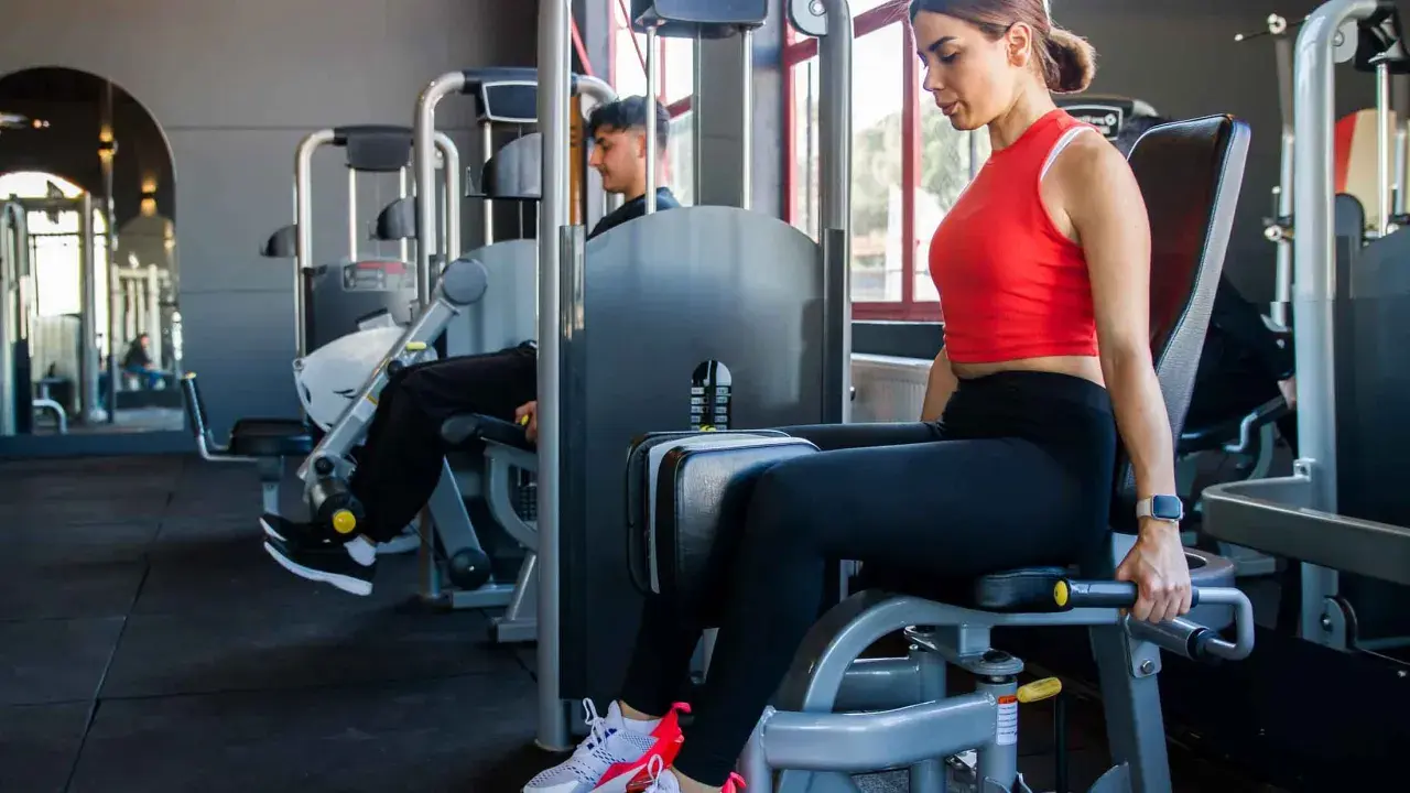 Frau trainiert ihre abduktoren muskeln an einem Gerät im Fitnessstudio. Ein Mann trainiert im Hintergrund.