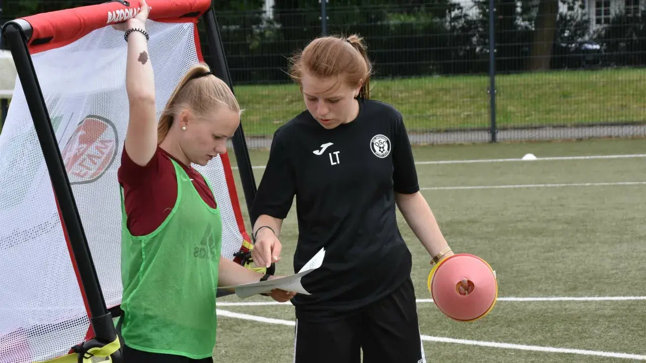 Zwei junge Fußballerinnen besprechen den Trainingsplan. Die eine hält einen Zettel, die andere einen Trainingskegel. Perfekte f jugend spielzeit.
