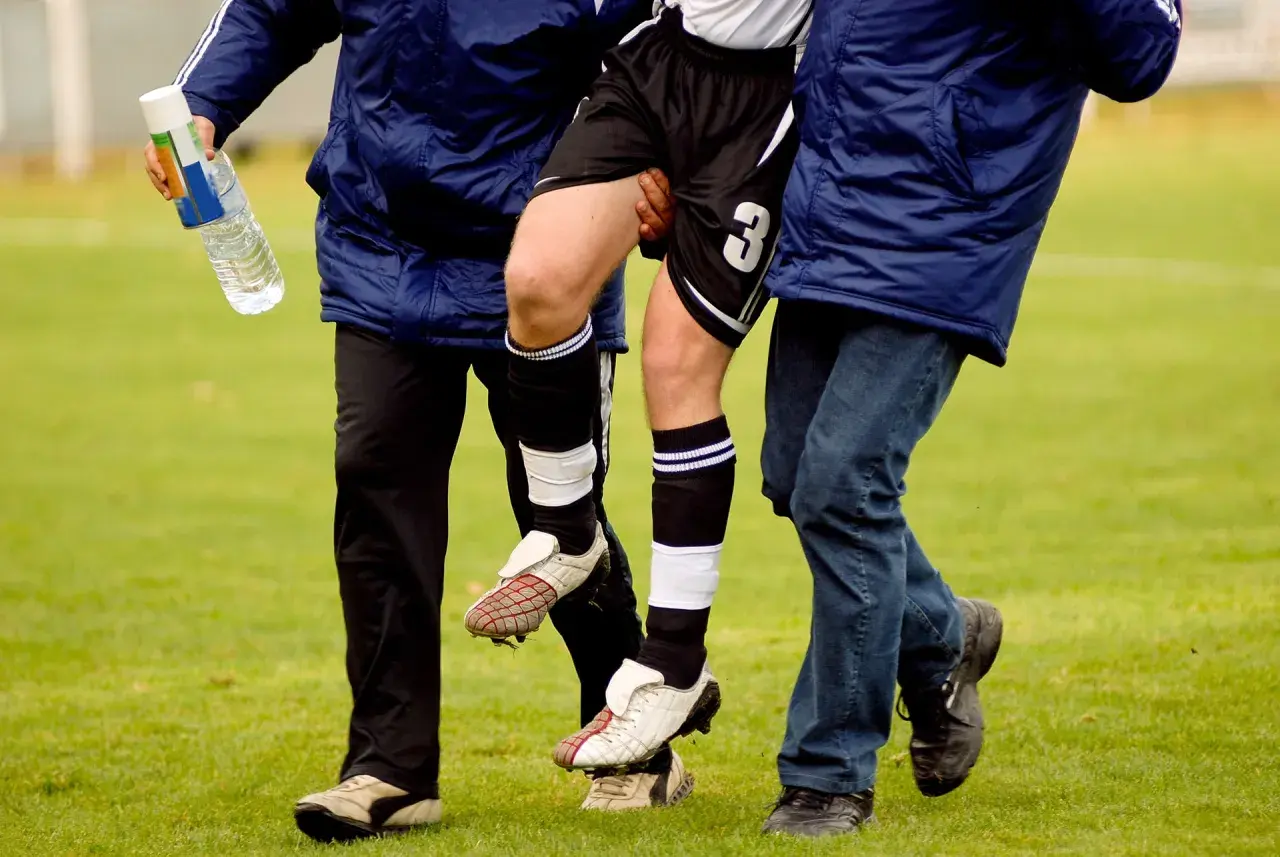 Fußballer mit verletztem Bein wird von zwei Helfern gestützt. Die Adduktoren-Gegenspieler sind hier wohl überlastet.