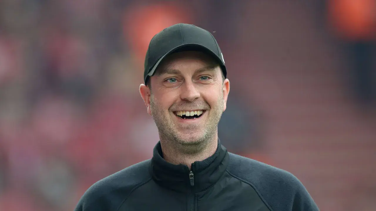 Lachender rb leipzig trainer mit schwarzer Kappe und Trainingsjacke vor unscharfem Publikum.