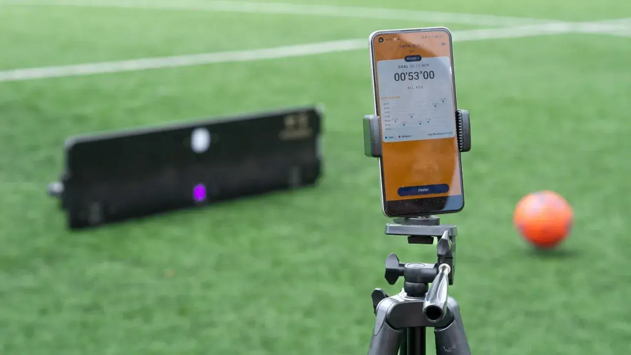 Smartphone mit Trainings-App für SAQ-Training auf einem Stativ, daneben ein Ball und ein Trainingsgerät auf einem Fußballfeld.