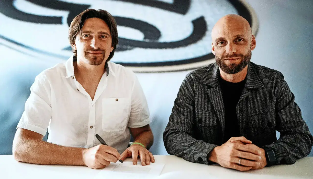 Zwei M&auml;nner unterschreiben einen Vertrag zwischen Verein und Spieler. Der eine Mann schreibt, der andere blickt in die Kamera.