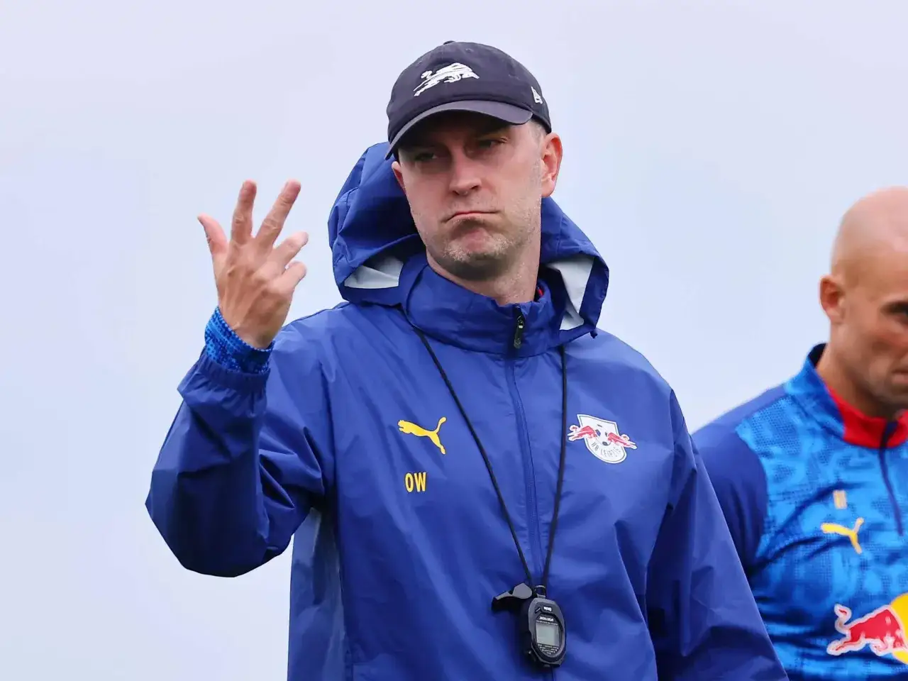 Ole Werner gestikuliert mit drei Fingern, trägt eine blaue Puma-Jacke mit dem RB Leipzig Logo und eine Kappe.