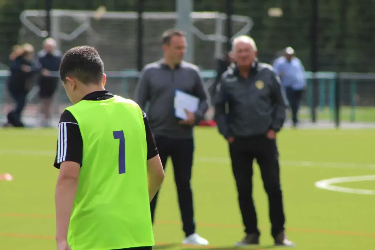 Ein junger Fußballer mit der Nummer 1 auf seinem neongrünen Trainingsleibchen wird von zwei Männern beobachtet, die wie ein Scout und Trainer aussehen.