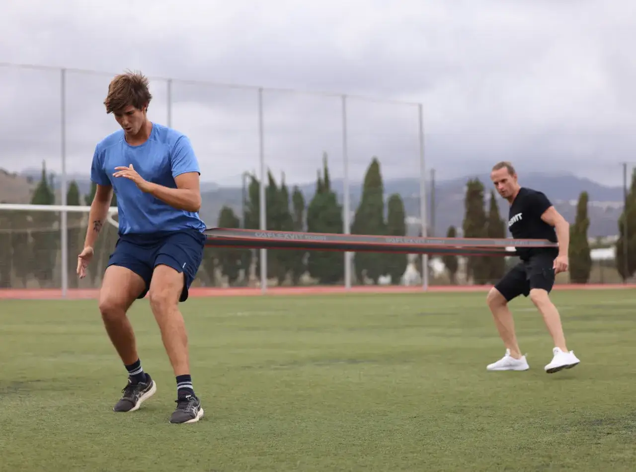 Zwei M&auml;nner trainieren mit einem Widerstandsband auf einem Fu&szlig;ballfeld. Der vordere Mann in Blau zieht, der hintere Mann in Schwarz h&auml;lt dagegen. Ein intensiver Aufbau Training.