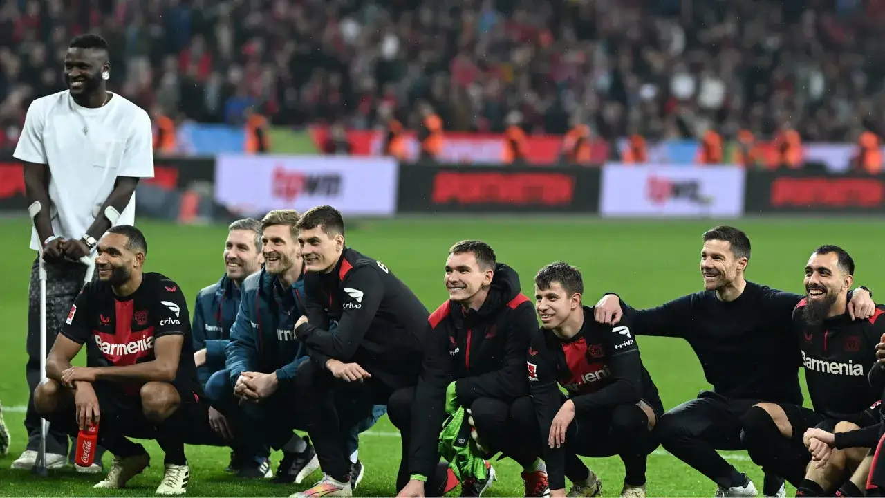 Xabi Alonso feiert mit dem Team von Leverkusen. Die Spieler sitzen auf dem Rasen, einige mit Kr&uuml;cken.