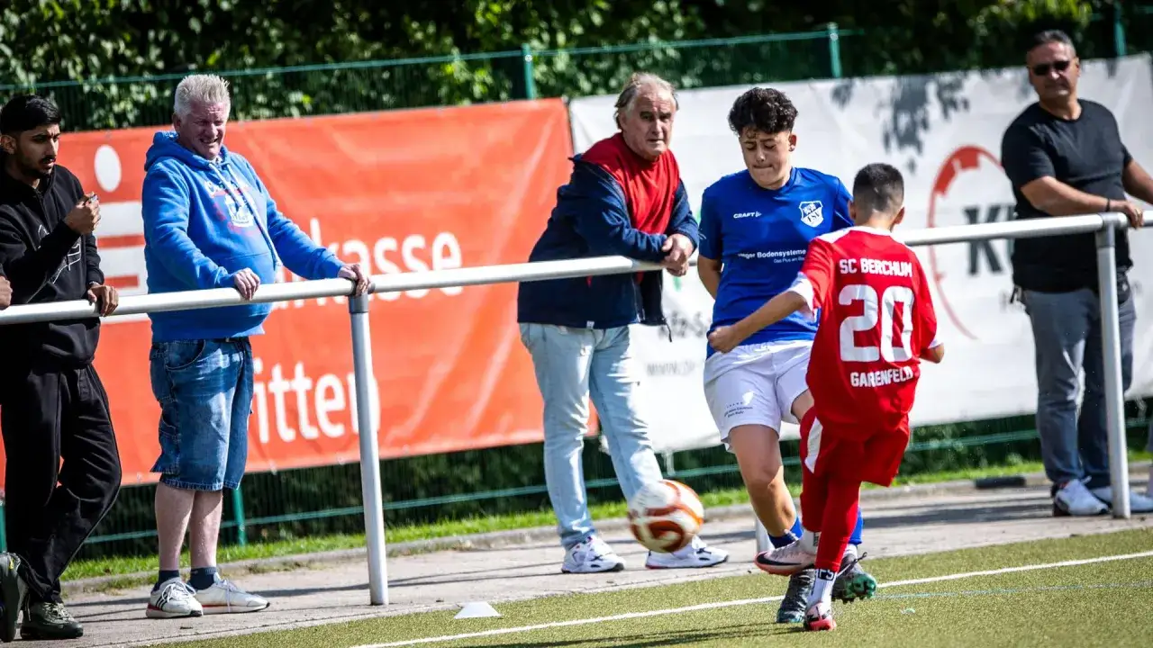 Fu&szlig;ball-Eltern schauen ihren Kindern beim Spiel zu. Ein Junge im roten Trikot mit der Nummer 20 kickt den Ball.