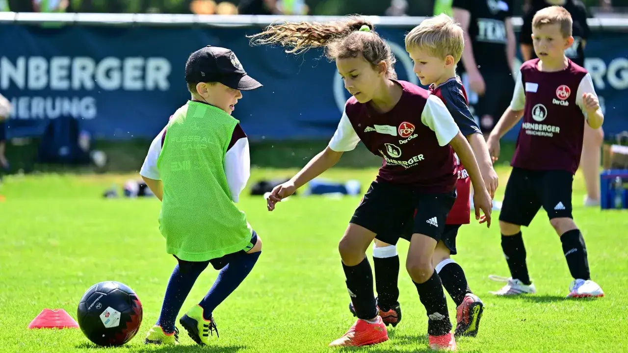 Kinder im Mannschaftssport Fu&szlig;ball &uuml;ben auf einem gr&uuml;nen Rasen. Ein Junge mit gr&uuml;ner Weste dribbelt den Ball, w&auml;hrend ein M&auml;dchen und ein weiterer Junge ihn beobachten.